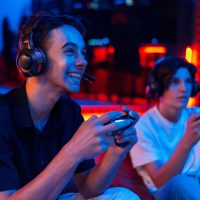 Two teen friends are playing a game console Two teen friends in headphones are playing a game console using gamepads and smiling while sitting on bean bags. Blue and red illumination