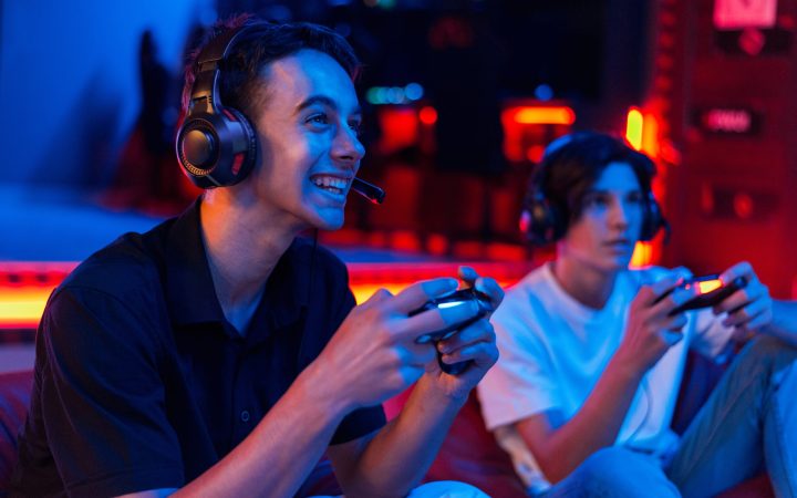 Two teen friends in headphones are playing a game console using gamepads and smiling while sitting on bean bags. Blue and red illumination
